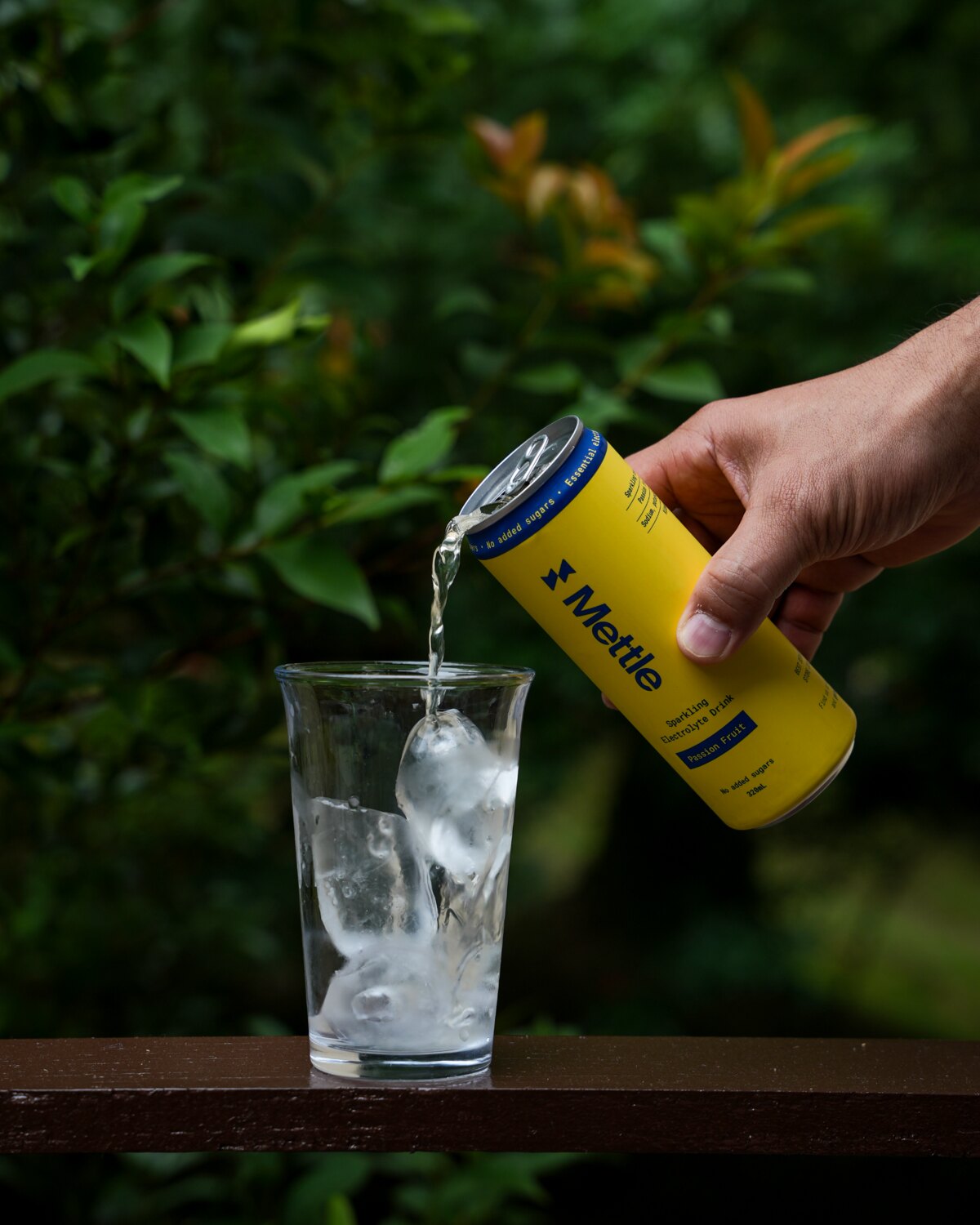 A hand pouring Passion Fruit Mettle into a glass of ice on a railing